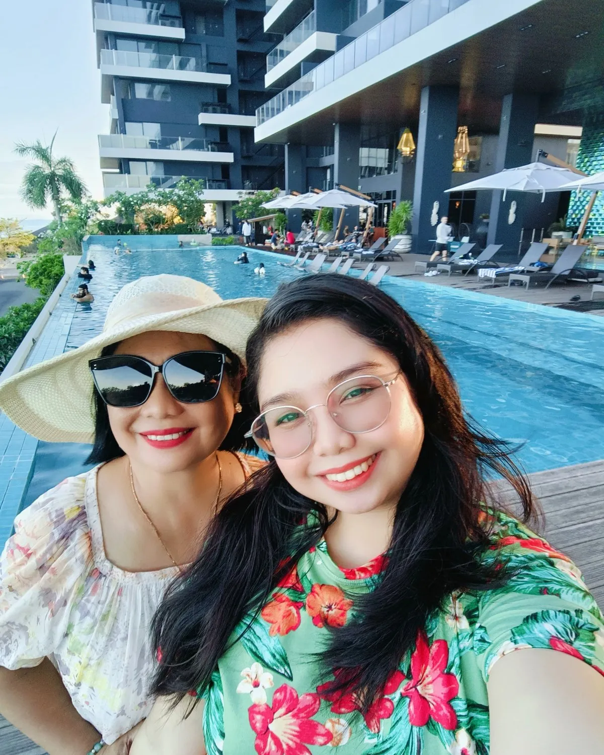 Beth Gucio and her daughter Angel smiling in a sunny selfie by a resort pool, with palm trees behind them
